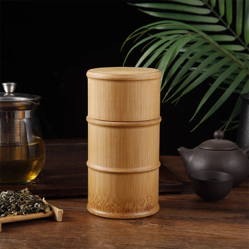 Bamboo tea canister on a wooden surface with tea-making items and green leaves in the background.